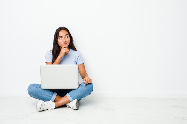 Mujer india de raza mixta joven sentado trabajando en la computadora portátil mirando hacia los lados con expresión dudosa y escéptica.