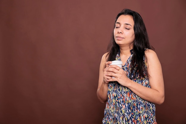 Mujer india relajada tomando café para llevar, disfrutando de capuchino caliente con los ojos cerrados. Persona sosteniendo una taza de papel de té, persona satisfecha con café con leche para llevar, tiro medio de estudio sobre fondo marrón