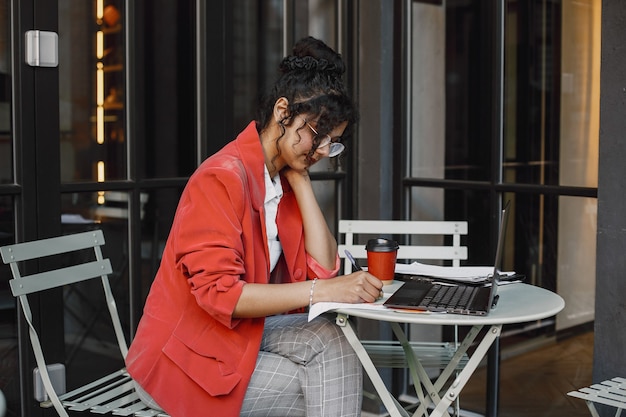 Mujer india trabajando en un portátil en un café de la calle. Usar ropa elegante e inteligente: chaqueta, gafas