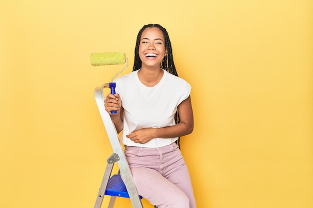 Foto mujer indonesia con rodillo en escalera en amarillo riendo y divirtiéndose