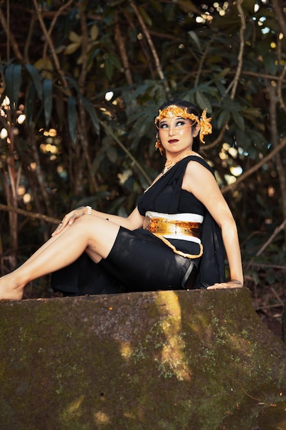 Mujer indonesia sentada en la roca con un traje de baile negro mientras usa una corona dorada y un collar dorado
