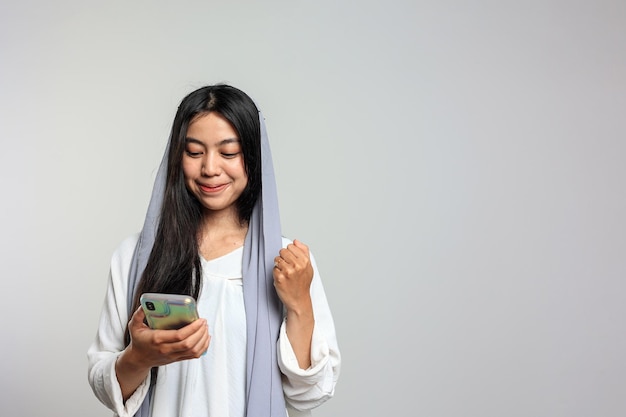 Mujer indonesia con velo feliz usando el teléfono móvil sonríe emocionada