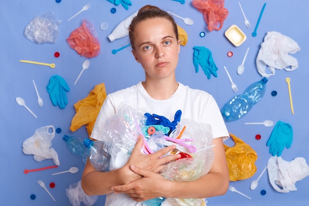 Una mujer infeliz con camisa blanca aislada en una pared azul con basura alrededor sosteniendo basura mirando a la cámara quiere salvar nuestro planeta y reducir la contaminación por desechos