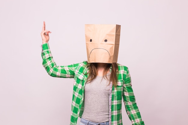 Foto mujer infeliz con emoticon triste delante de la bolsa de papel en la cabeza sobre fondo blanco.