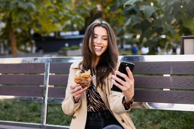 Una mujer influyente atractiva sostiene un teléfono celular y un croissant en sus manos graba un video o toma una foto Una mujer joven está sentada en un banco en la calle