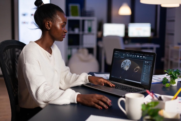 Mujer ingeniera afroamericana enfocada que trabaja en el prototipo de engranajes industriales a altas horas de la noche en la oficina de la empresa de inicio. Idea de construcción de productos de ingeniería en la pantalla del portátil