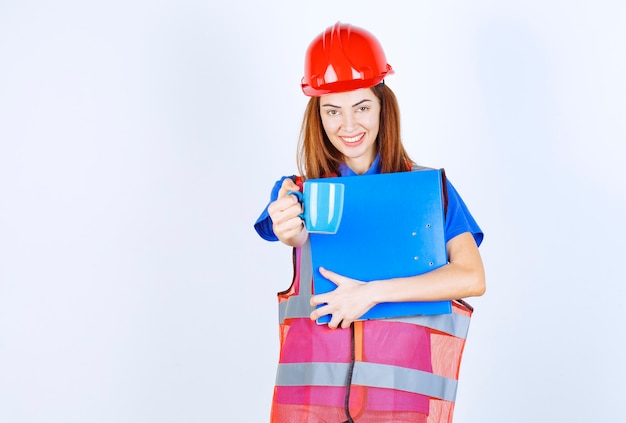 Mujer ingeniera en casco rojo sosteniendo una carpeta azul y ofreciendo una taza de bebida a su colega.