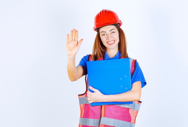 Mujer ingeniera en casco rojo sosteniendo una carpeta de proyecto azul y deteniendo algo.