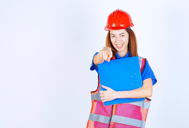 Mujer ingeniera en casco rojo sosteniendo una carpeta de proyecto azul y presentando a alguien o algo.