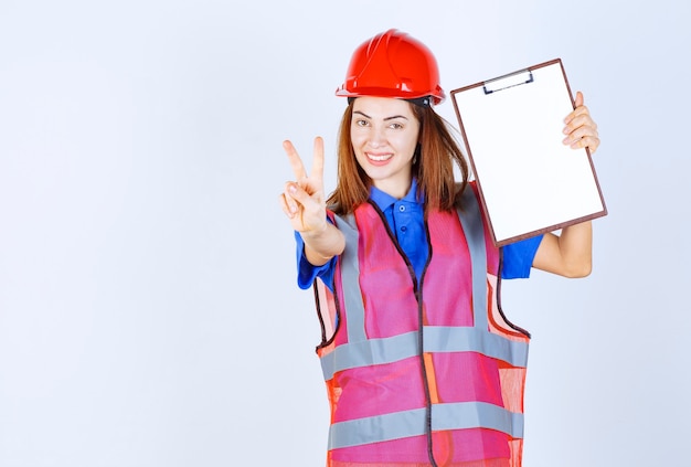 Mujer ingeniera en uniforme sosteniendo un archivo de informes en blanco y mostrando el signo de satisfacción.