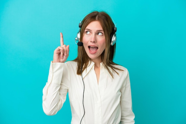 Mujer inglesa de telemarketing que trabaja con un auricular aislado en un fondo azul con la intención de darse cuenta de la solución mientras levanta un dedo