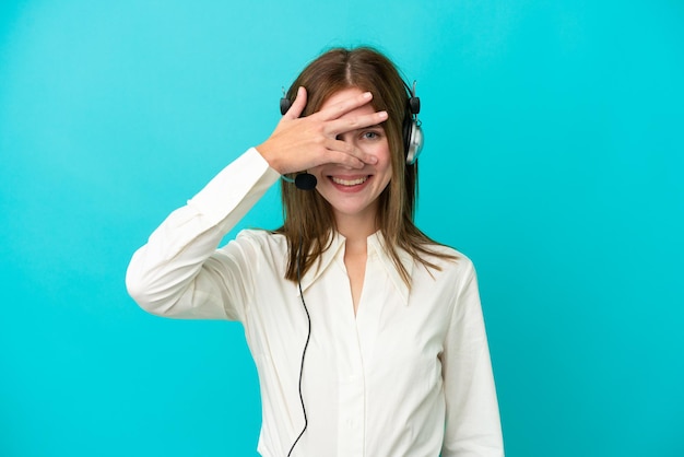 Mujer inglesa de telemarketing que trabaja con un auricular aislado de fondo azul que cubre los ojos con las manos y sonríe