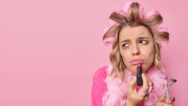 Mujer insatisfecha aplica lápiz labial sostiene pestañas curlerrr mira tristemente hacia otro lado usa rodillos para el cabello ha filtrado maquillaje después de llorar aislado sobre fondo rosa espacio en blanco para su promoción