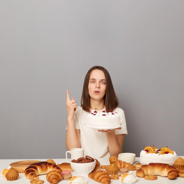 Mujer inspirada con camiseta blanca aislada sobre fondo gris con comida trampa sosteniendo pastel levantó el dedo con la idea de señalar el área de publicidad