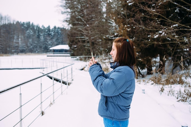 Mujer en invierno con teléfono toma fotos de la naturaleza en el parque en un día frío