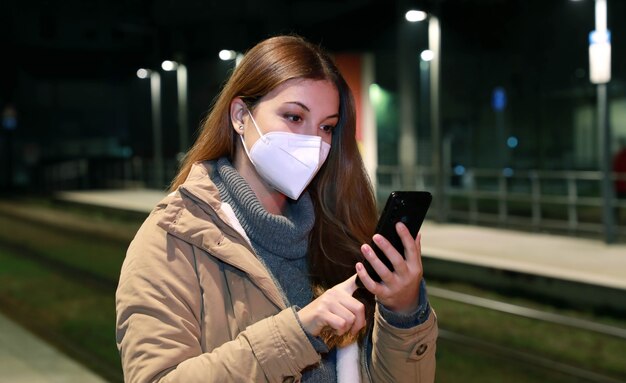 Mujer de invierno vistiendo máscara protectora KN95 FFP2 con smartphone espera el tren en la estación vacía por la noche