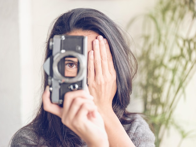 Mujer irreconocible con cabello largo y oscuro mirando a través de una cámara fotográfica de película retro mientras cubre media cara con la mano y toma fotos