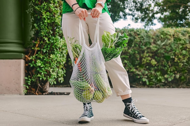 Mujer irreconocible con camisa verde sosteniendo una bolsa de malla con verduras de pie en la calle
