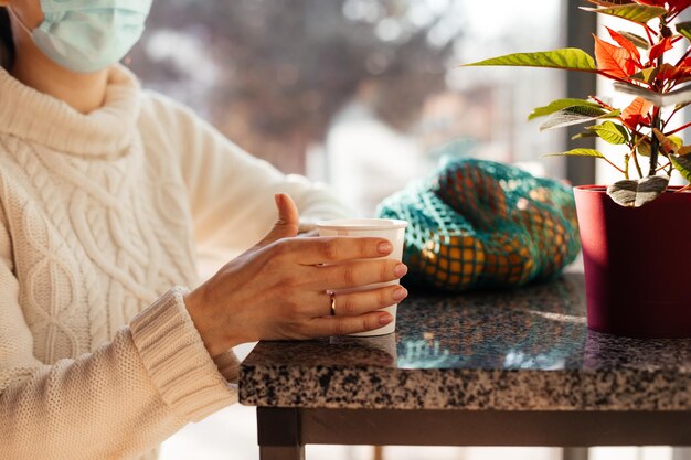 Mujer irreconocible con máscara protectora sentada cerca de la ventana sosteniendo una taza de café de papel Primer plano de la mano de una mujer joven calentándose con una taza de té caliente