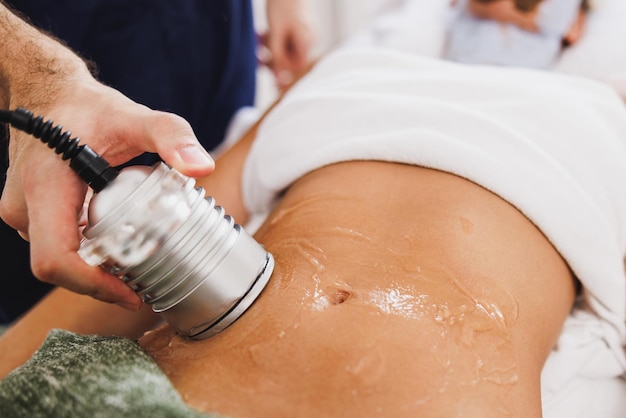 Mujer irreconocible recibiendo un tratamiento de cavitación por ultrasonido para reducción de grasa en el abdomen en el salón de belleza.