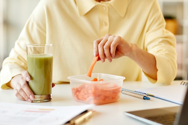 Mujer irreconocible sentada en el escritorio en la oficina comiendo zanahoria fresca y bebiendo batido de verduras h