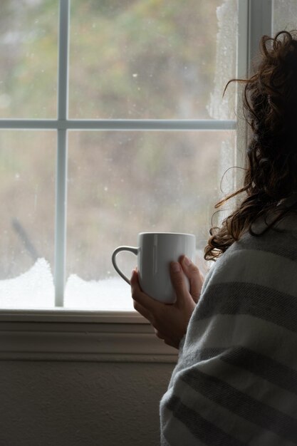 mujer irreconocible sosteniendo café frente a la ventana con nieve