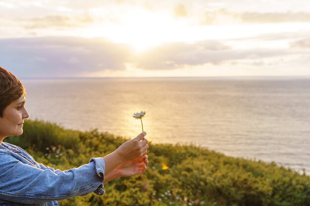 Foto mujer irreconocible sosteniendo una flor en un acantilado al atardecer. vista horizontal de la mujer caucásica viajando y haciendo turismo al aire libre sosteniendo un diente de león.