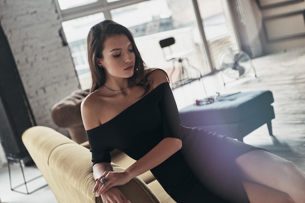 Mujer irresistible. Vista superior de una mujer joven y atractiva en un elegante vestido negro con una hendidura profunda simplemente descansando mientras está sentado en el sofá
