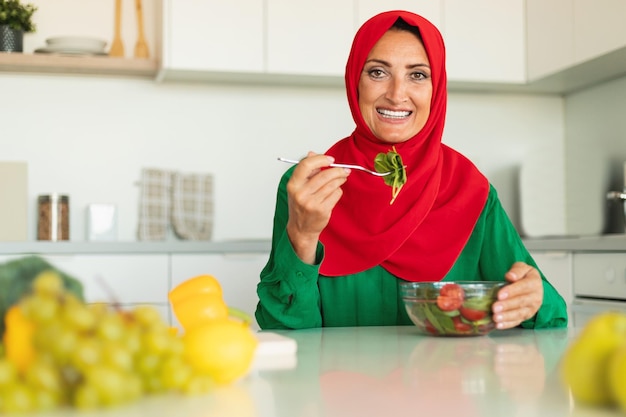 Mujer islámica madura feliz comiendo ensalada de verduras frescas para la cena sentada en la cocina y sonriendo a la cámara