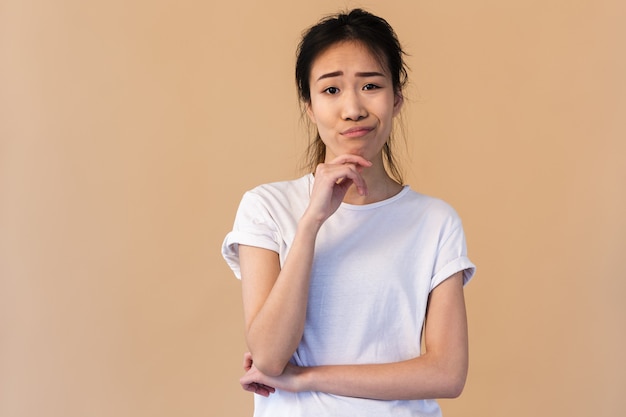 Mujer japonesa disgustada vistiendo camiseta básica tocando la barbilla y haciendo pucheros aislado sobre pared beige en studio