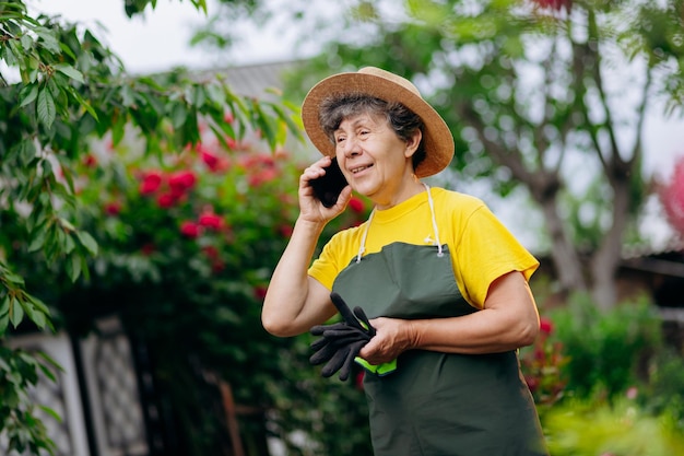 Mujer jardinera senior en un sombrero trabajando en su jardín y habla por teléfono El concepto de jardinería creciendo y cuidando flores y plantas