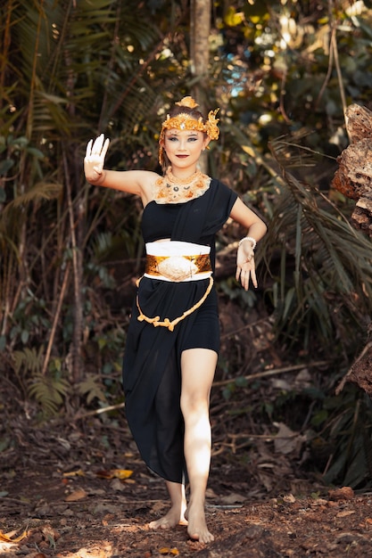 Mujer javanesa bailando en una camiseta negra y falda negra con corona dorada y accesorios dorados en su cuerpo dentro de la jungla