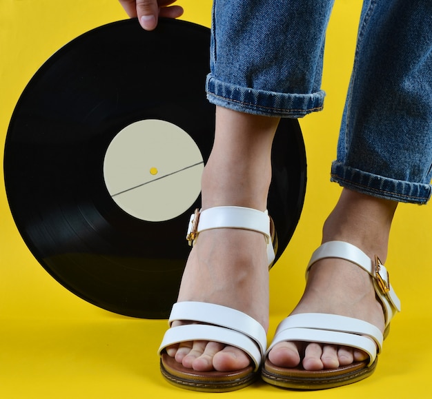 Mujer con jeans y sandalias está sosteniendo una placa de vinilo retro en una pared amarilla