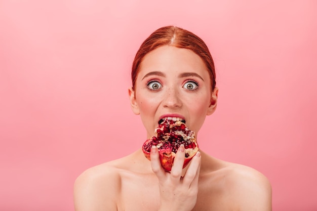 Mujer de jengibre asombrada comiendo granate Vista frontal de una chica atractiva con granada madura aislada sobre fondo rosa