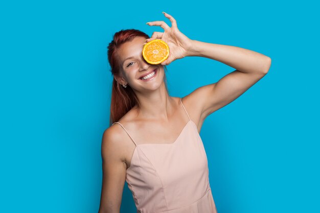 La mujer de jengibre está sonriendo mientras se cubre el ojo con una naranja posando sobre una pared azul