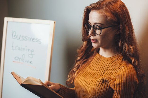 Mujer de jengibre con gafas está leyendo un libro durante las clases de negocios en línea