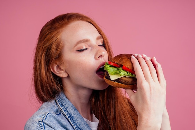 Mujer de jengibre pelirroja disfrutando de una gran chuleta de hamburguesa y usando una chaqueta de jeans americana demin en el concepto tradicional de estudio de fondo rosa de EE.UU.