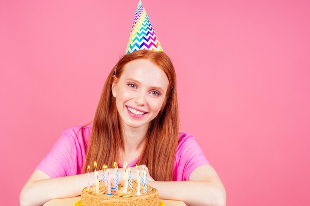 Mujer de jengibre pelirroja pidiendo un deseo soñando y soplando velas en el pastel con cono de sombrero de cumpleaños en el fondo rosa del estudio