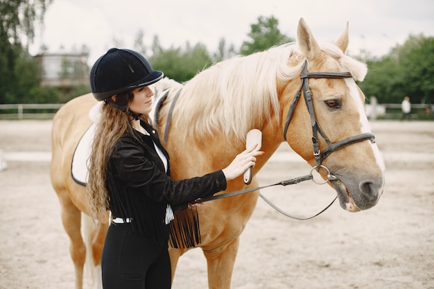 Mujer jinete peina su caballo en un rancho. La mujer tiene cabello largo y ropa negra. Ecuestre femenina tocando su caballo marrón.