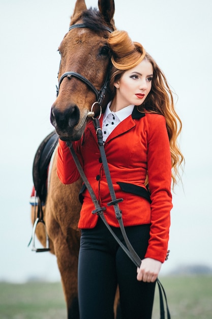 Mujer jockey pelirroja en un cárdigan rojo y botas altas negras con un caballo a caminar