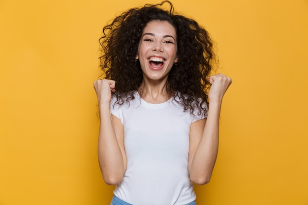 Foto mujer joven de 20 años con el pelo rizado gritando y apretando los puños aislados en amarillo