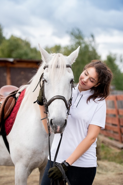 Mujer joven abrazando a un caballo de carreras blanco y mirándola mientras se relaja en un entorno rural