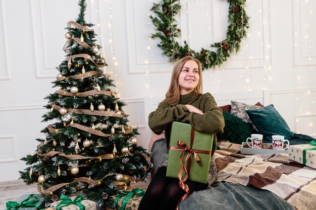 Una mujer joven abre regalos de Navidad en un ambiente de Año Nuevo y se regocija La mujer está en un dormitorio luminoso y acogedor bellamente decorado para Navidad