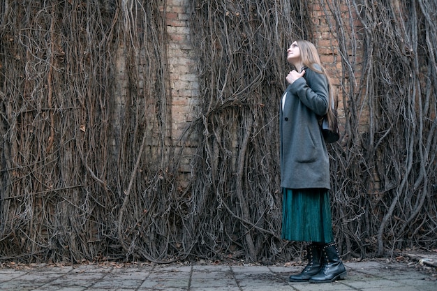Mujer joven con un abrigo se encuentra en el contexto de una pared de hiedra marchita. Vista lateral.