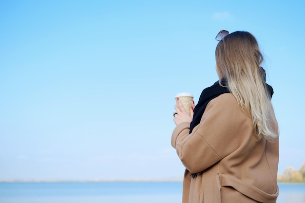 Una mujer joven con un abrigo marrón y una bufanda negra se para con una taza de café en las manos y disfruta del cielo azul y la vista al mar