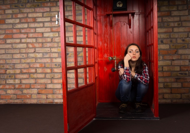 Mujer joven aburrida esperando una llamada en una cabina telefónica pública sentada en el suelo con una expresión de mal humor