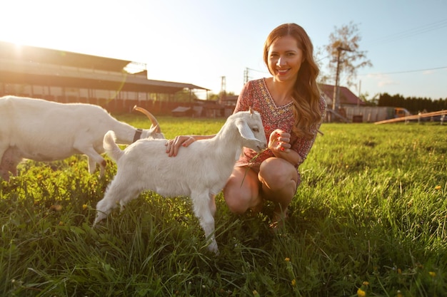 Mujer joven acariciando cabrito, sonriendo, alimentándolo con hierba, el fuerte sol de la tarde brilla sobre el fondo de la granja