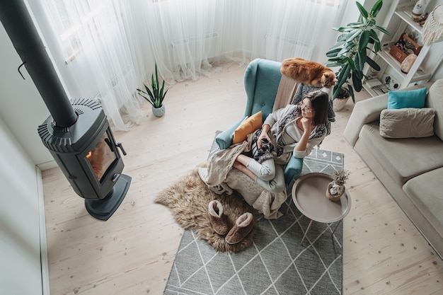 Mujer joven en un acogedor sillón y un plaid sentado junto a la chimenea con un gato doméstico, bebiendo té en casa