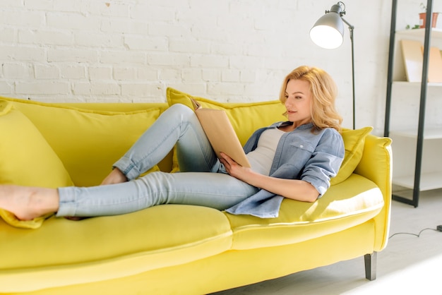 Mujer joven acostada en un acogedor sofá amarillo y leyendo un libro