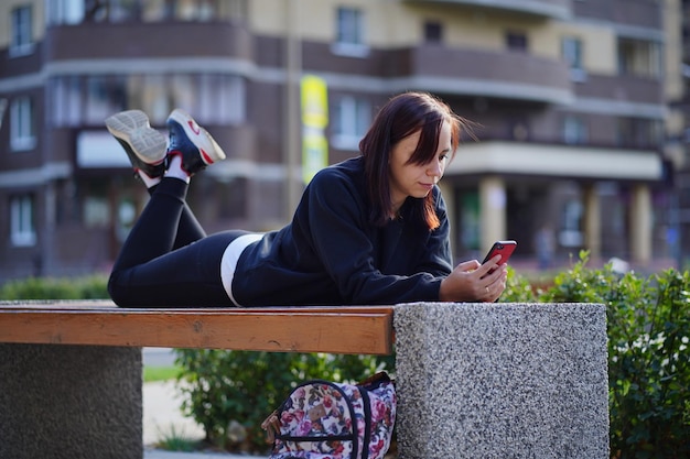 Mujer joven acostada boca abajo afuera en un banco con las piernas levantadas y navegando por su teléfono celular Una mujer que usa un teléfono inteligente usa Internet en el patio de un edificio residencial